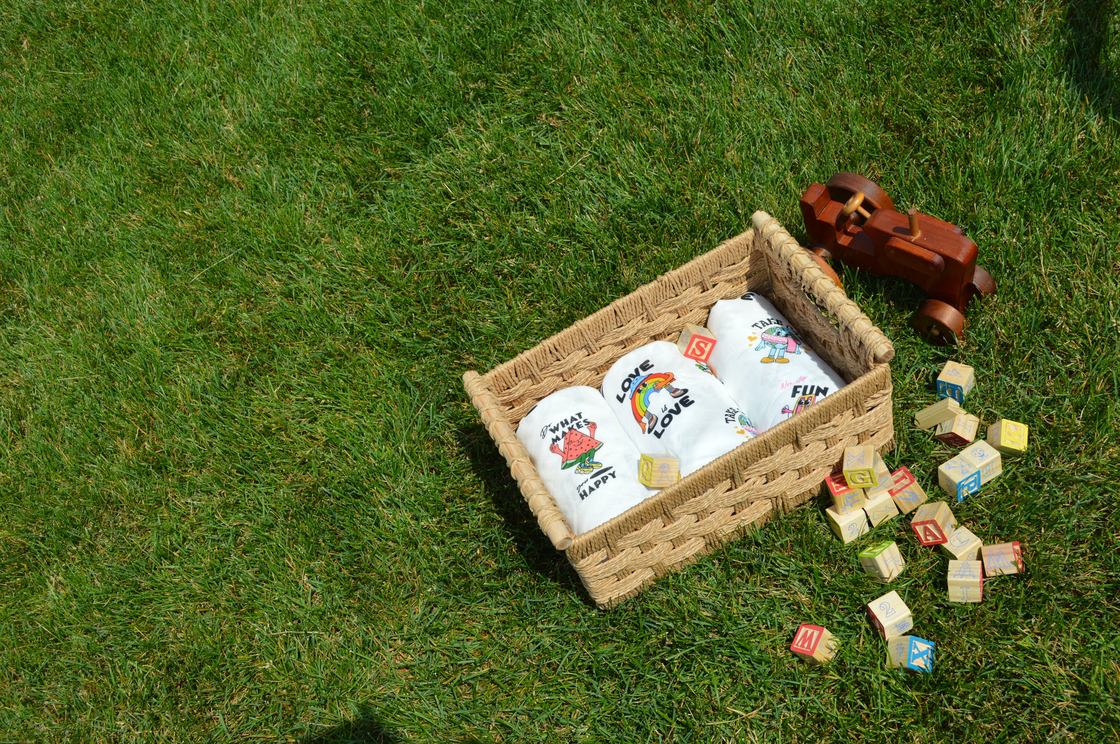 Wicker basket with baby items on grass, including a toy and colorful blocks. | Muslinco